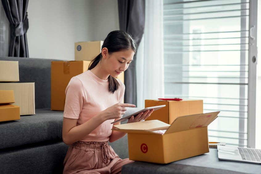 A lady sits around shipping boxes using a tablet to run her online selling business